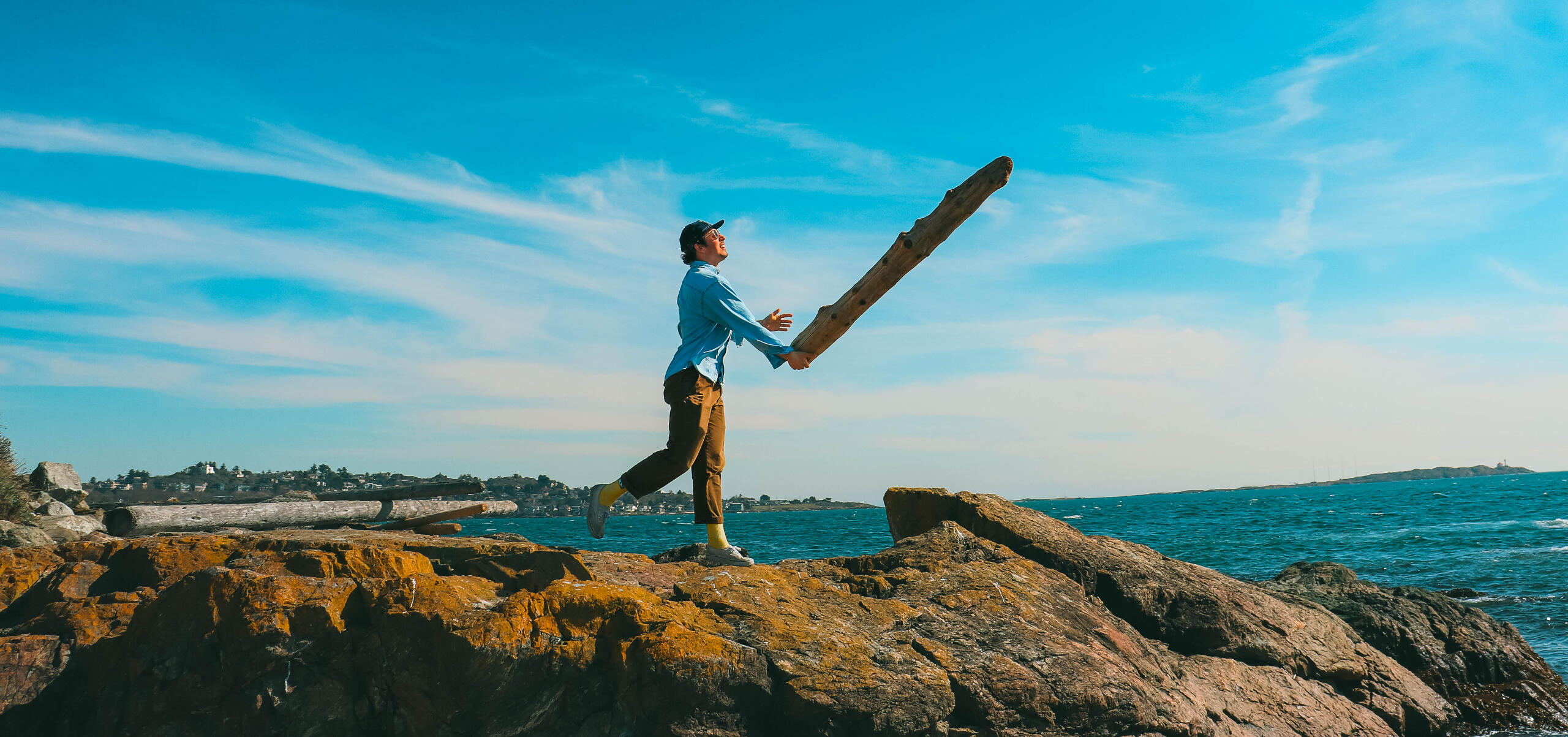 Throw Your Ideas Out There A portrait and landscape photo of a man in colorful clothing walking on rocky terrain throwing a large log upwards with the ocean, Victoria British Columbia and blue sky in the background.