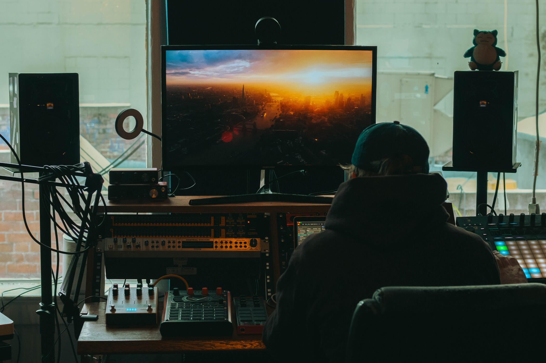 Vault 22 Man seated at a sound mixing desk in a music studio downtown Kelowna, facing a large monitor displaying a cityscape at sunset. The room has various audio equipment and speakers.