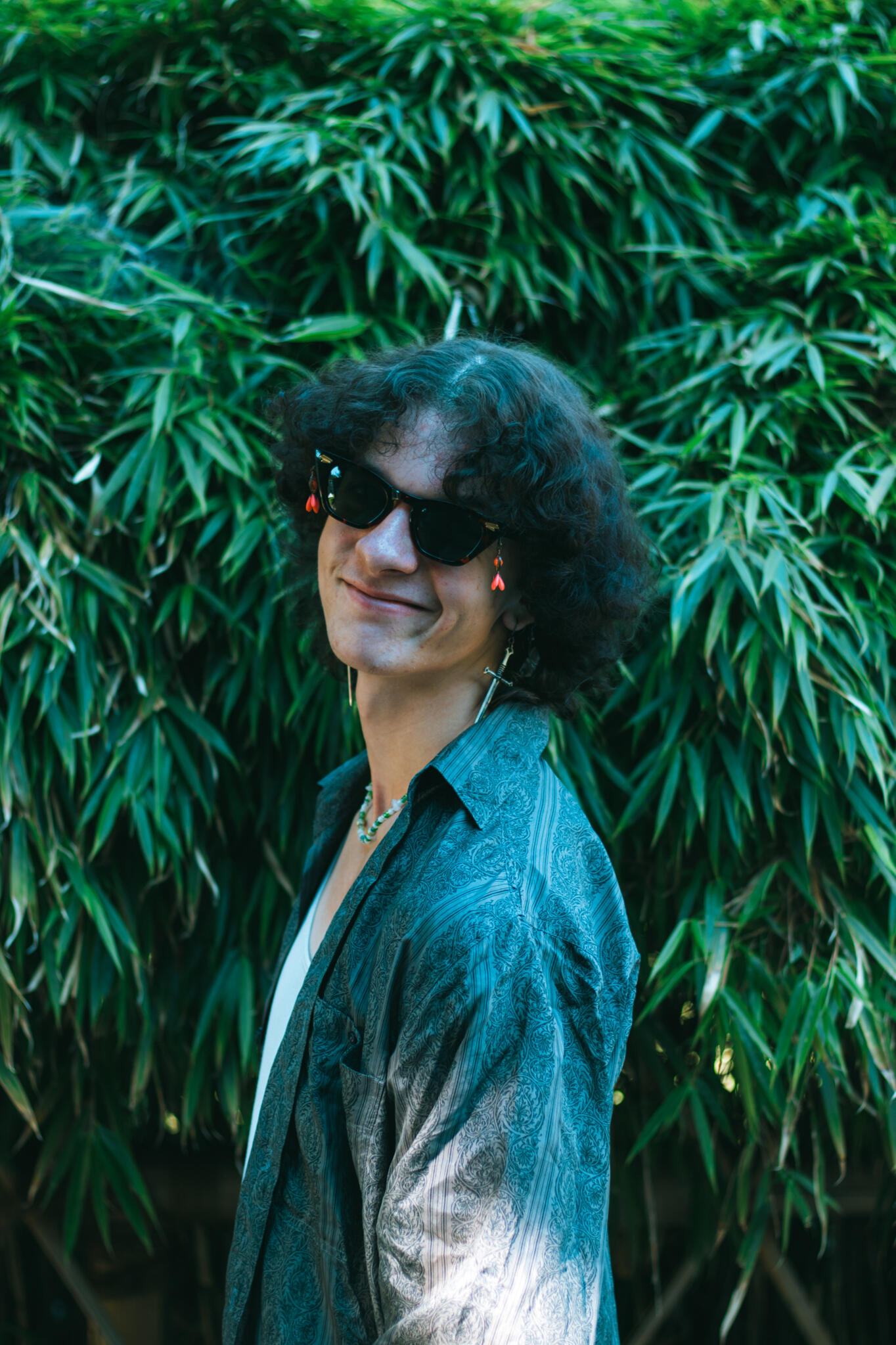 Kasugai Gardens A man wearing sunglasses and with curly hair poses in front of a vibrant green bush, showcasing a cheerful demeanor.