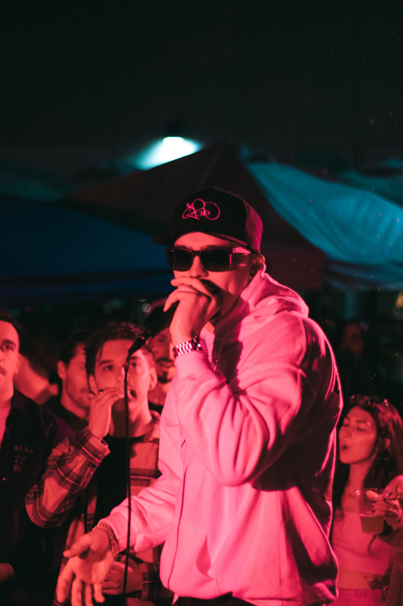Never Ending Summer A man in a white hoodie illuminated by pink lighting and wearing sunglasses performs in front of a large audience in the rain while singing into a microphone.